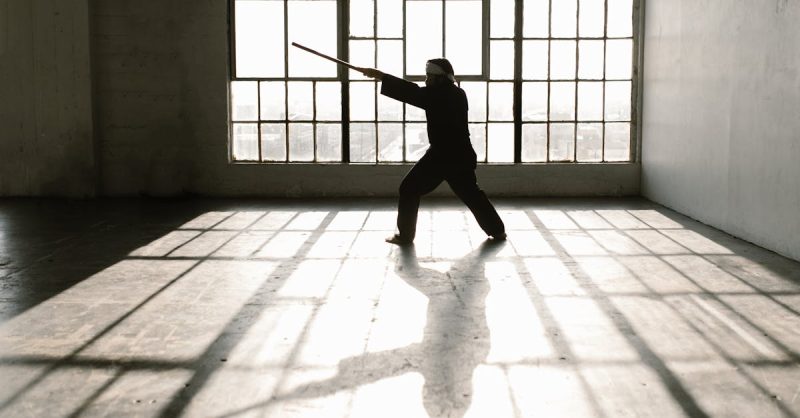 Silhouette of martial artist practicing with a staff in a sunlit studio.