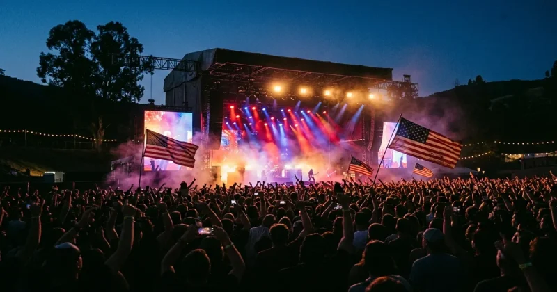 Massive outdoor concert crowd at dusk with American flags and dramatic stage lighting at a rock festival
