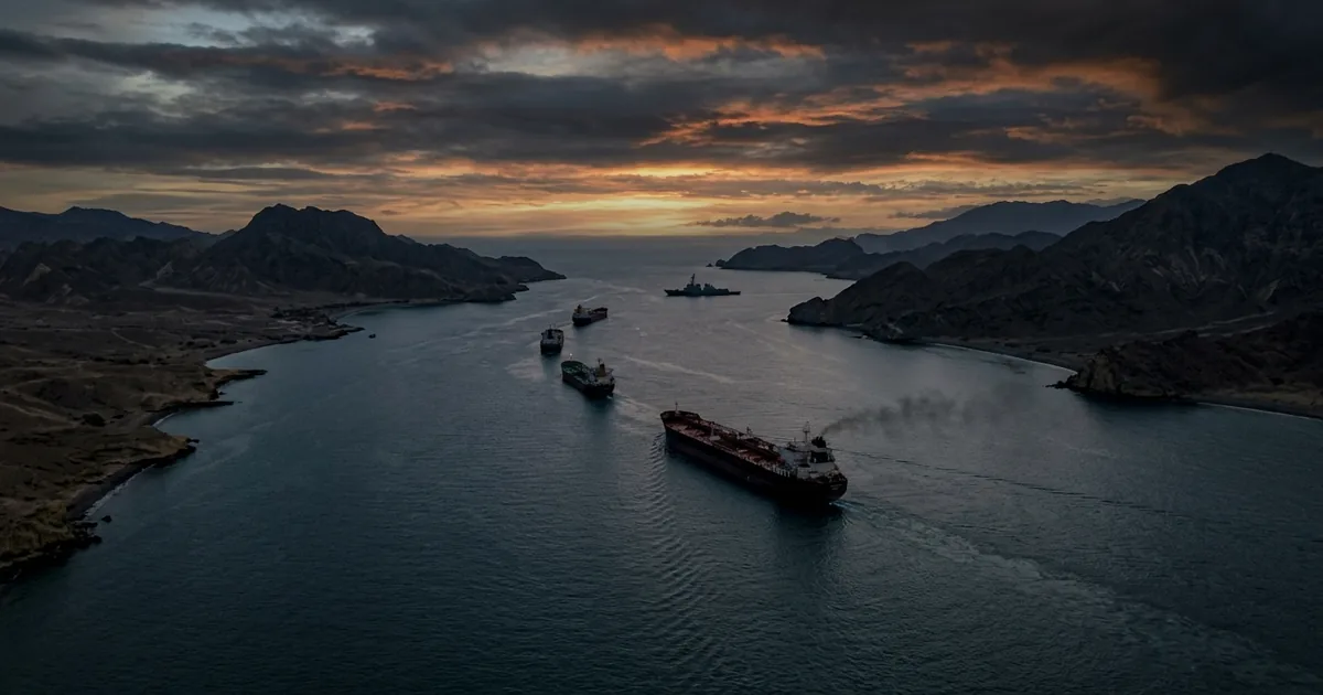 Oil tankers and a distant naval warship on the Strait of Hormuz at dusk under a stormy sky