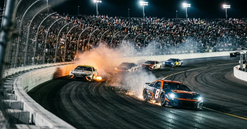 Stock cars racing through a banked turn at night under stadium lights with sparks flying