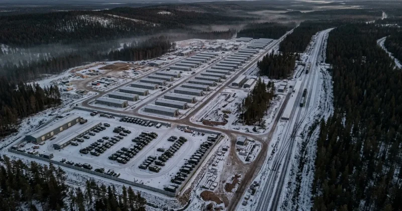 Aerial view of rows of new gray barracks, a fenced motor pool of military vehicles, and fresh rail sidings in a snowy northern forest at dusk