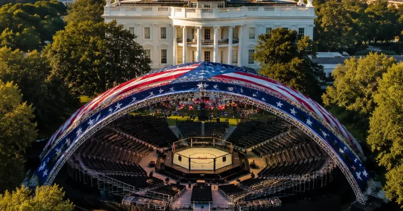 Aerial view of a red, white and blue arched canopy arena with a UFC octagon on the White House South Lawn, the White House facade rising behind it at golden hour