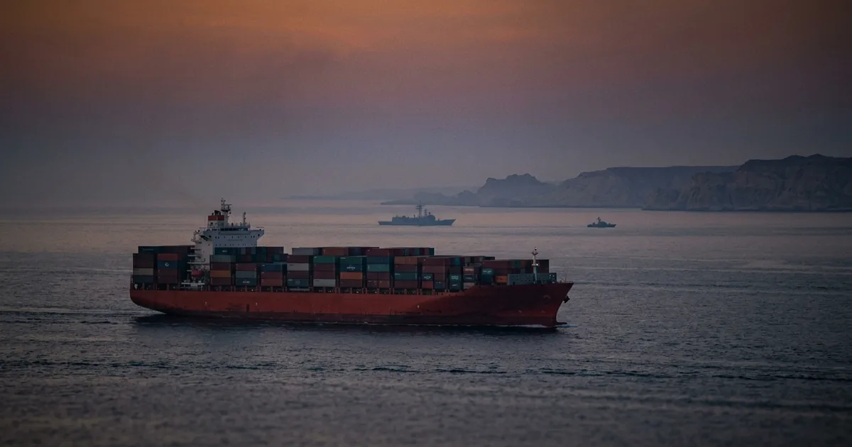A commercial cargo container ship transits the Strait of Hormuz at dusk as a gray naval warship and a small fast patrol boat sit on the hazy horizon