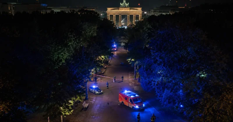 Emergency lights illuminating Berlin Tiergarten park at night with Brandenburg Gate in background after vehicle attack at Pride parade