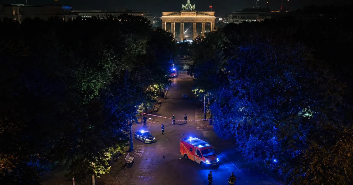 Emergency lights illuminating Berlin Tiergarten park at night with Brandenburg Gate in background after vehicle attack at Pride parade