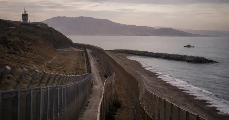 Twin barbed-wire border fences run down a hillside to a beach at Ceuta, with a rock breakwater extending into the Mediterranean and a patrol boat offshore