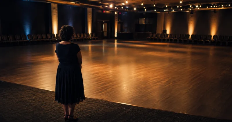 A lone person standing at the edge of a large empty dance floor in a dimly lit hall