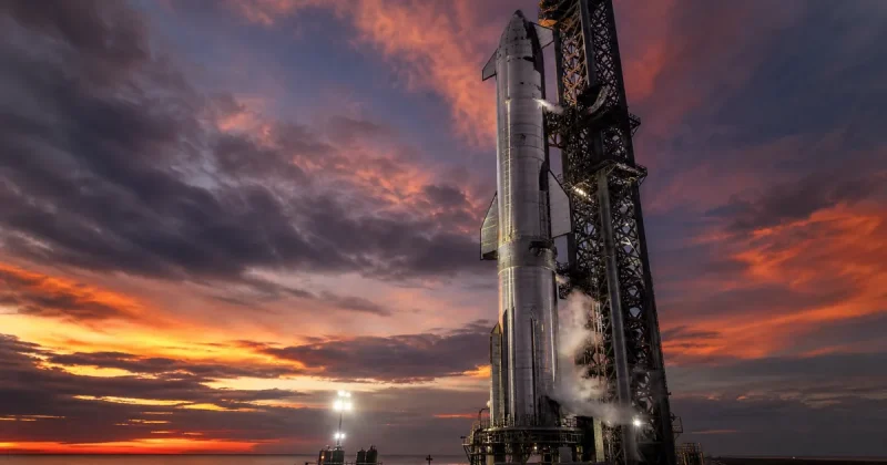 A massive stainless steel rocket on a launch pad at dusk with spotlights and vapor from fuel lines
