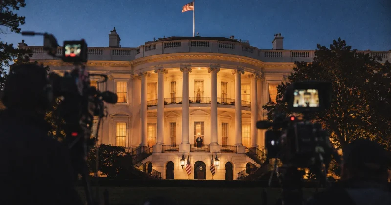 The White House at dusk with broadcast cameras positioned outside, American flags flanking the colonnade