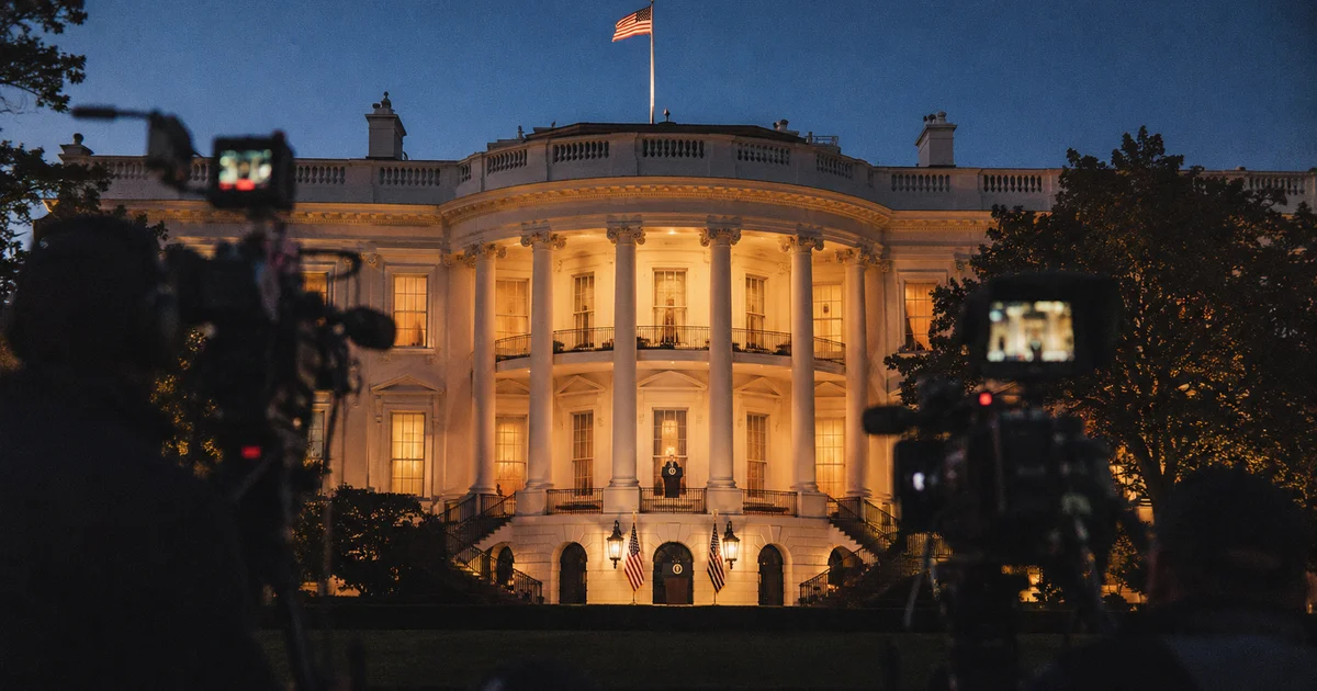 The White House at dusk with broadcast cameras positioned outside, American flags flanking the colonnade