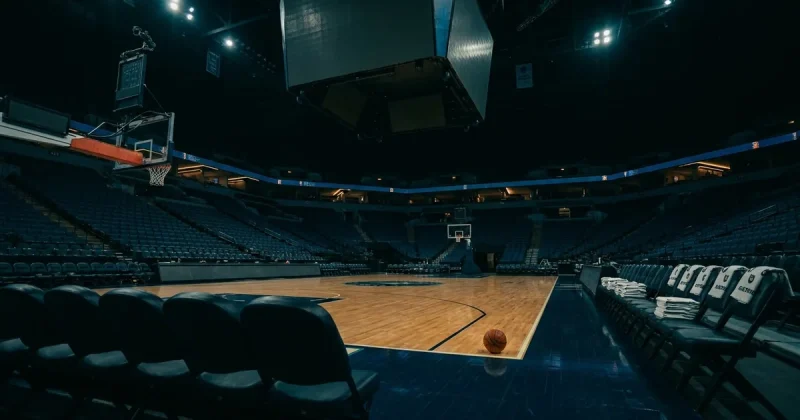 A dim empty basketball arena with a single orange ball on the hardwood, folded towels on the bench and dark scoreboard screens overhead