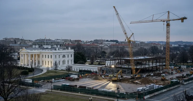 Tower cranes and steel framing rise over the cleared East Wing site beside the intact White House residence, behind green construction fencing