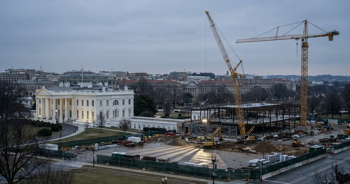 Tower cranes and steel framing rise over the cleared East Wing site beside the intact White House residence, behind green construction fencing