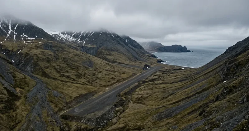 A narrow gravel runway running along the slope of a treeless tundra mountain on the Bering Sea coast, with high ridges on both sides and low cloud covering the summit