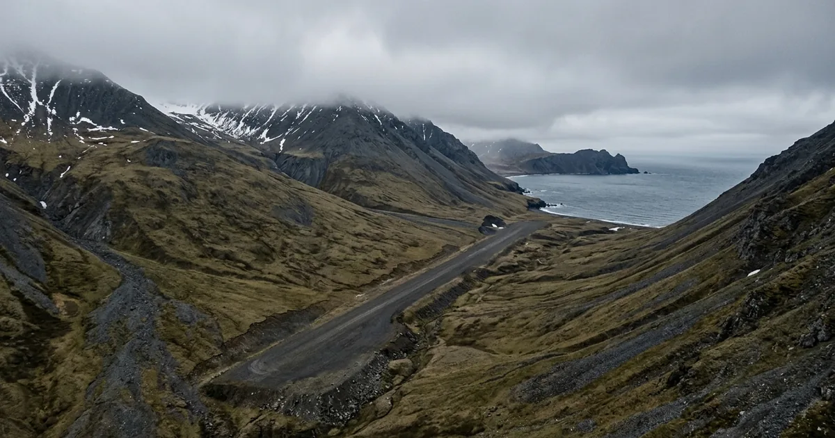 A narrow gravel runway running along the slope of a treeless tundra mountain on the Bering Sea coast, with high ridges on both sides and low cloud covering the summit