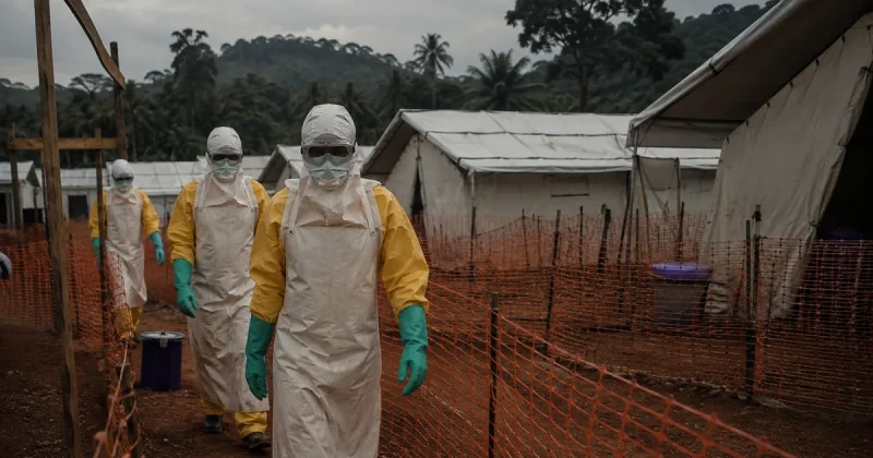 Health workers in full protective equipment, yellow coveralls, white aprons, goggles and green gloves, walk a fenced circuit between white canvas tents at an Ebola treatment centre