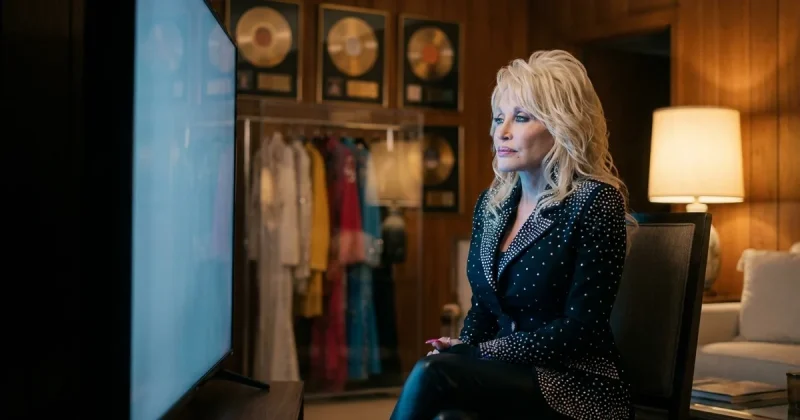 Dolly Parton seated alone in a wood-panelled Nashville room, lit by a large screen on one side and a lamp on the other, with framed gold records and a rack of stage costumes behind her