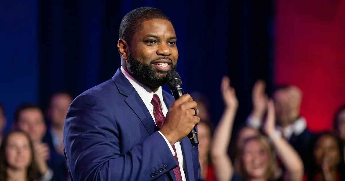A Black congressman in a navy suit speaks into a handheld microphone at a podium as an out-of-focus crowd applauds behind him