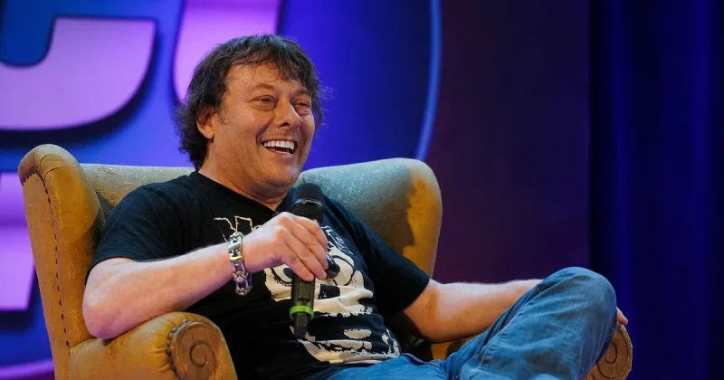 A man in his late forties with shoulder-length dark hair sits back laughing in an armchair on a convention stage holding a microphone, lit by warm key light against purple and blue stage lighting