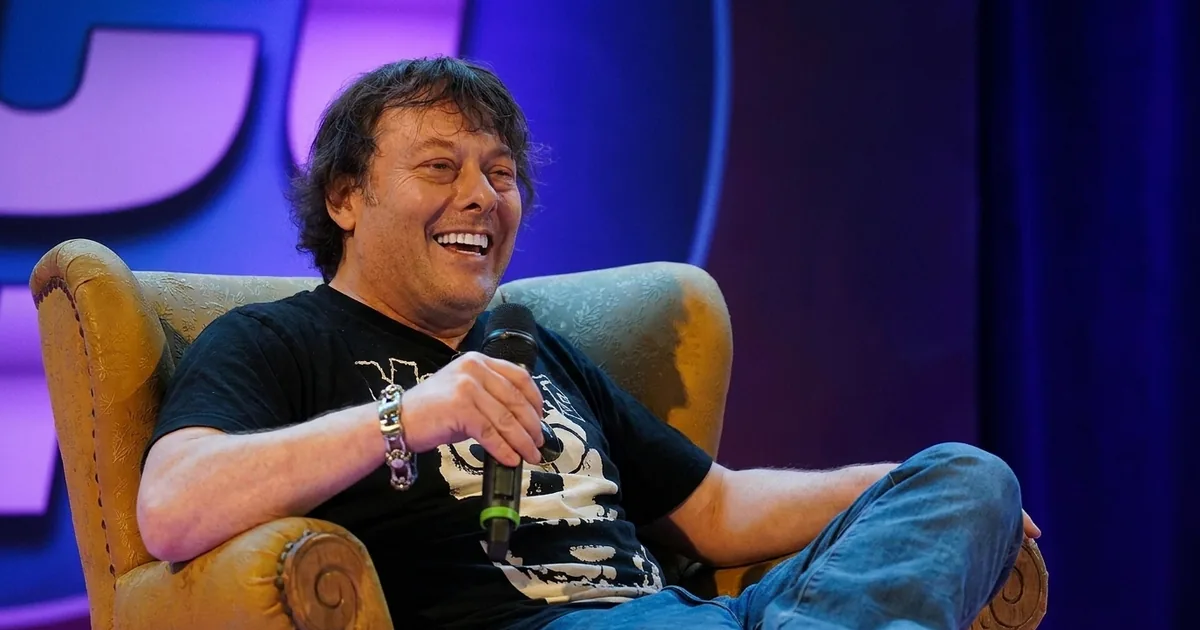 A man in his late forties with shoulder-length dark hair sits back laughing in an armchair on a convention stage holding a microphone, lit by warm key light against purple and blue stage lighting