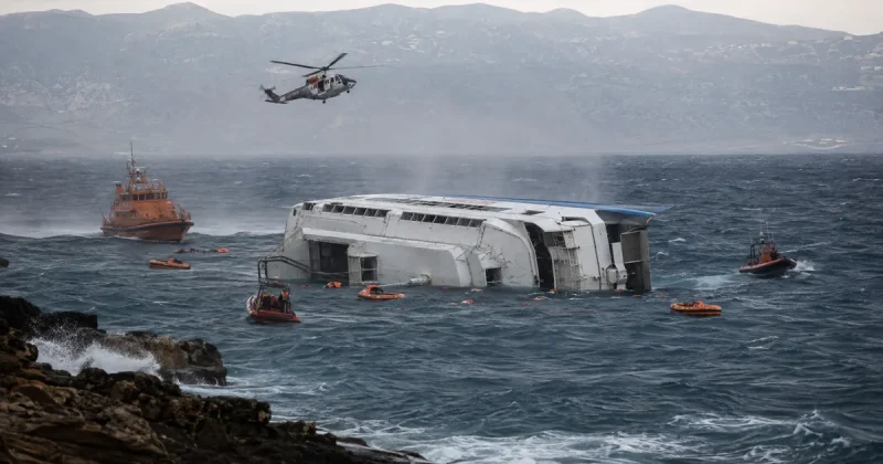 A white passenger catamaran lies capsized on its side in heavy grey seas while an orange coast guard vessel, rescue boats and a helicopter search among scattered life rafts near a rocky coastline.