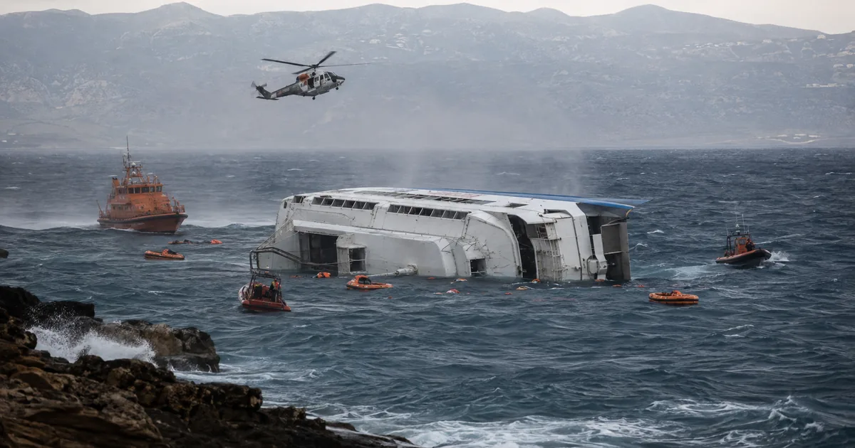 A white passenger catamaran lies capsized on its side in heavy grey seas while an orange coast guard vessel, rescue boats and a helicopter search among scattered life rafts near a rocky coastline.
