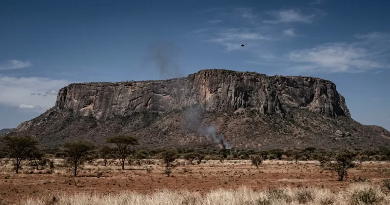 The flat-topped granite massif of Mount Ololokwe rises above dry red scrubland and acacia trees, with a thin column of grey smoke at the base of its cliffs and a distant helicopter overhead