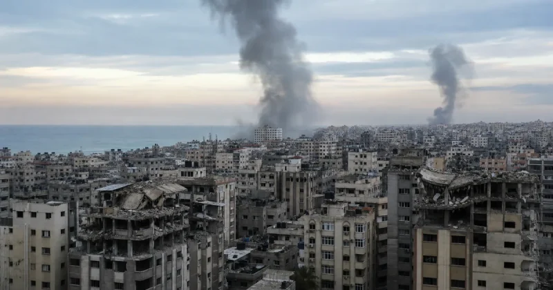 Two columns of smoke rise over a dense coastal skyline of damaged mid-rise concrete apartment blocks with collapsed upper floors, seen from above at dawn