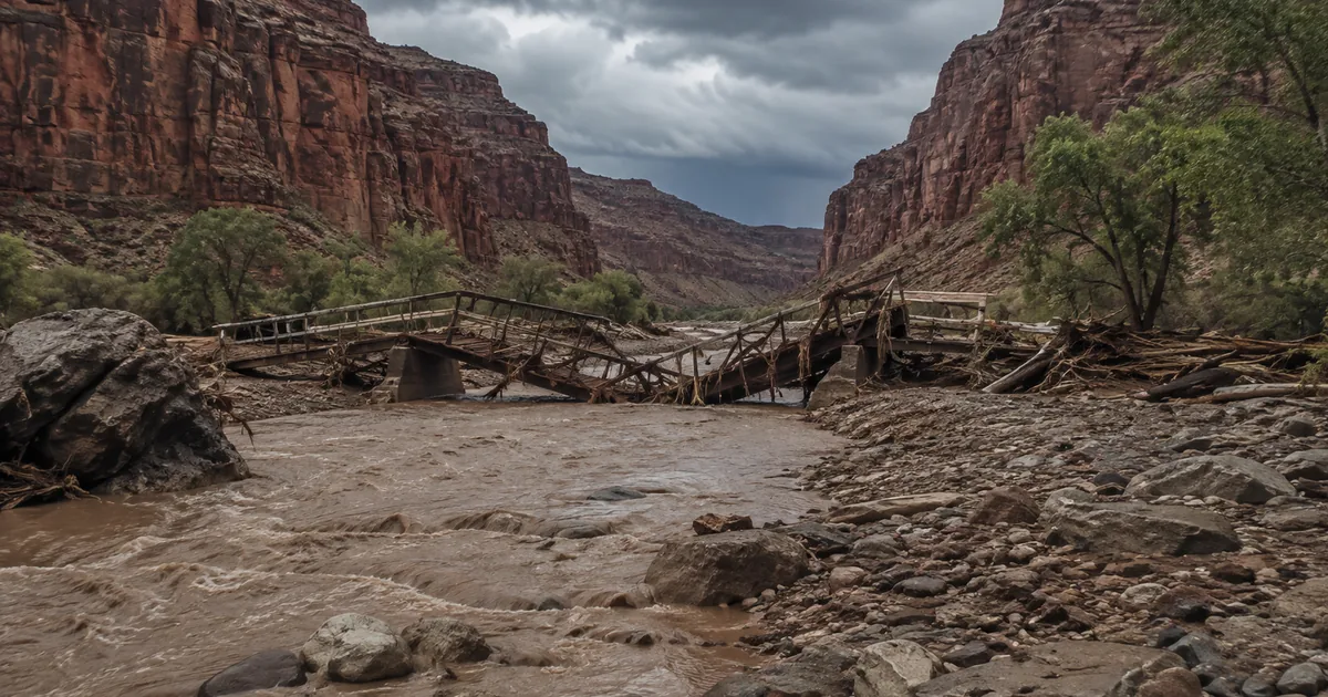 A collapsed timber and steel footbridge lies twisted across a silt-brown creek choked with boulders and flood debris, between steep layered canyon walls under storm clouds.