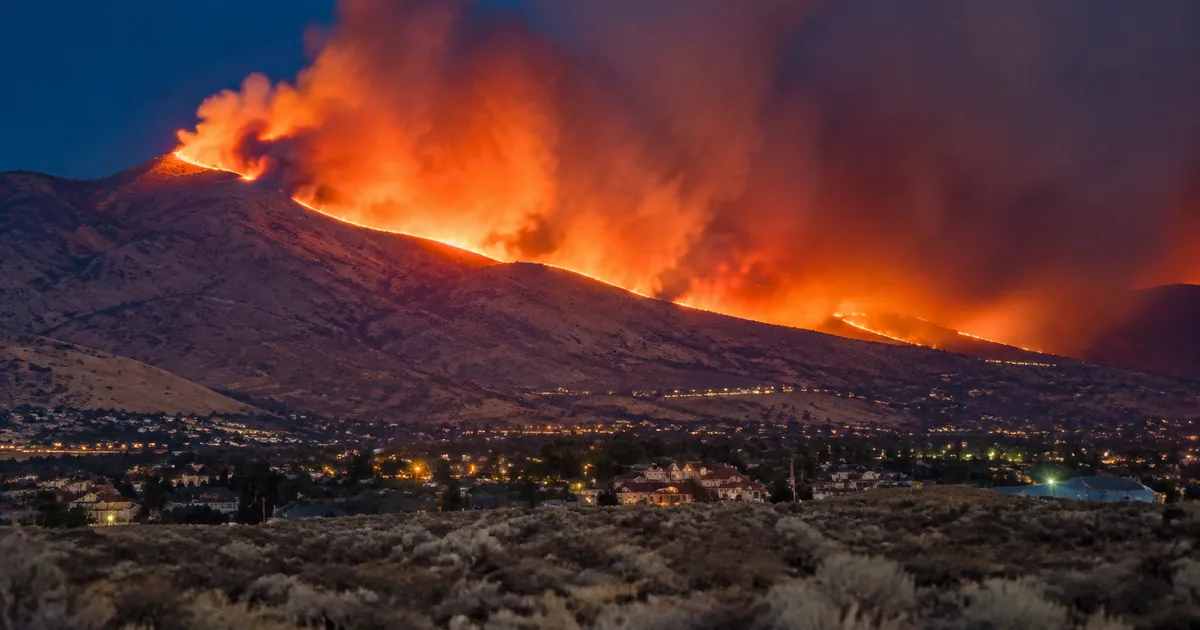 A wildfire burns along the ridgeline of a dry high desert mountain at dusk, throwing a glowing orange smoke column into a deep blue sky, with suburban homes and streetlights spread across the valley floor at the base of the slope.