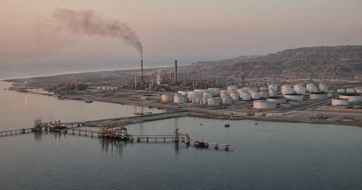 Aerial view at dawn of a coastal oil refinery beside calm water, with distillation columns, a field of white storage tanks, marine loading jetties, and a column of dark smoke rising from one stack