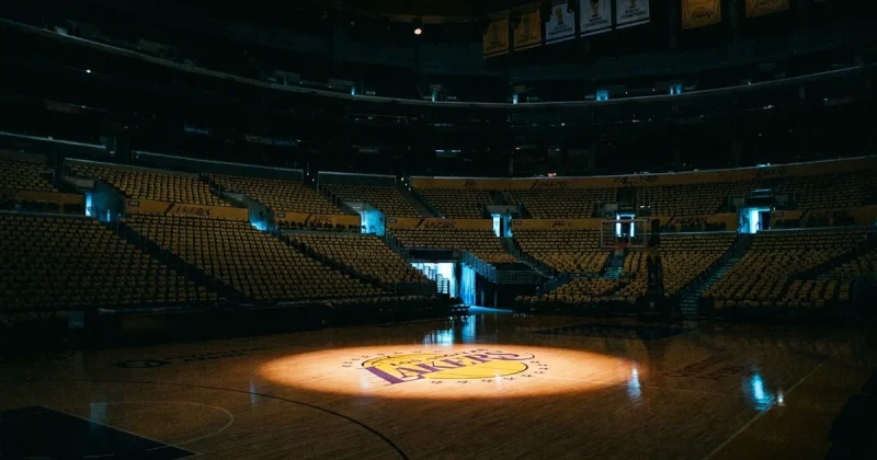 The darkened Los Angeles Lakers home court, empty gold and purple seats around it, with the center-court Lakers logo lit by one overhead spotlight