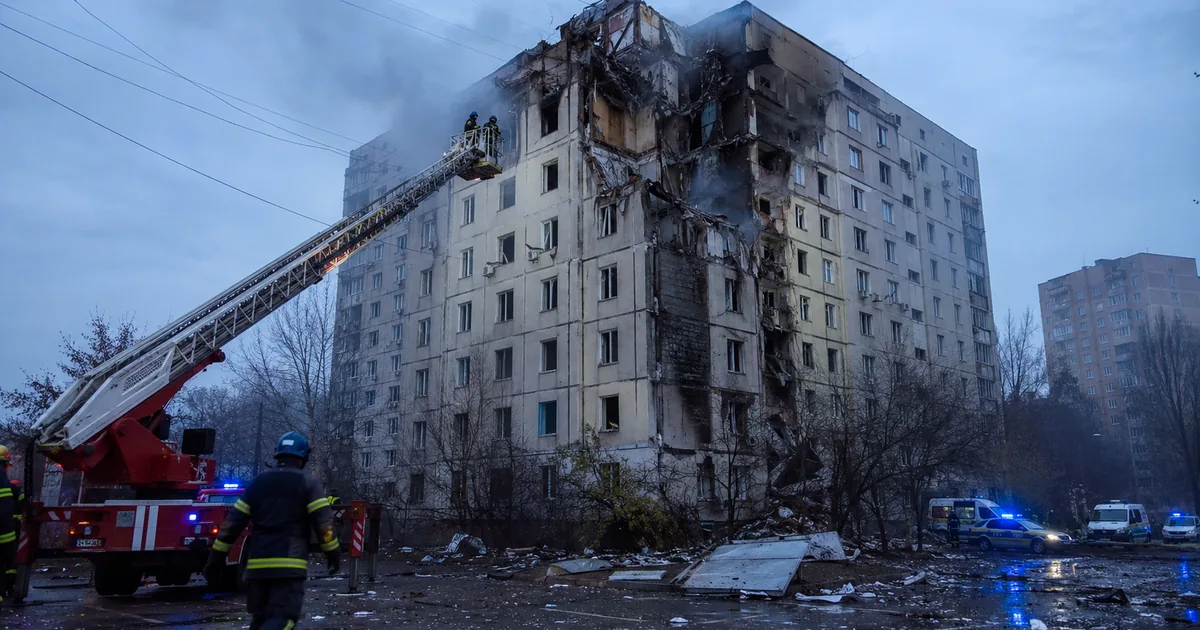 Firefighters on an extended red aerial ladder work at the gutted upper floors of a nine-storey pale concrete apartment block, its facade blackened and torn open, with debris across wet asphalt and emergency vehicles at a cordon in the grey morning light