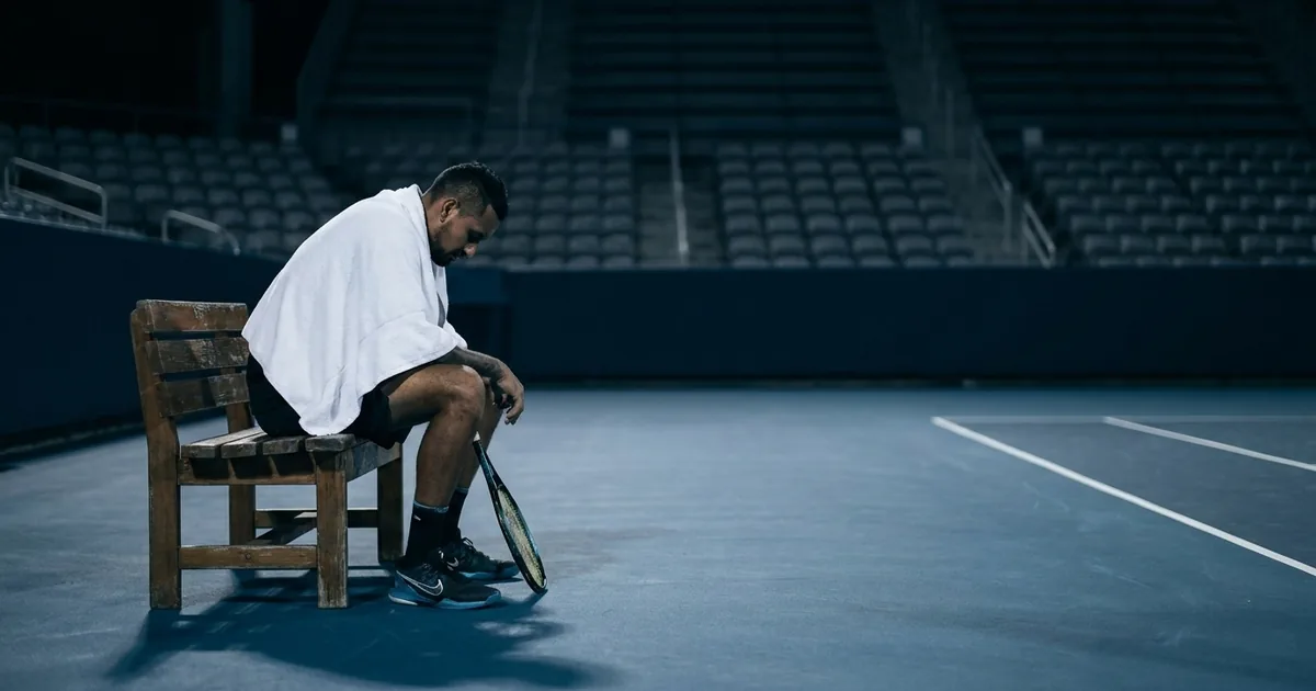 A dark-haired tennis player sits head-down on a courtside bench with a towel over his shoulder in a completely empty stadium