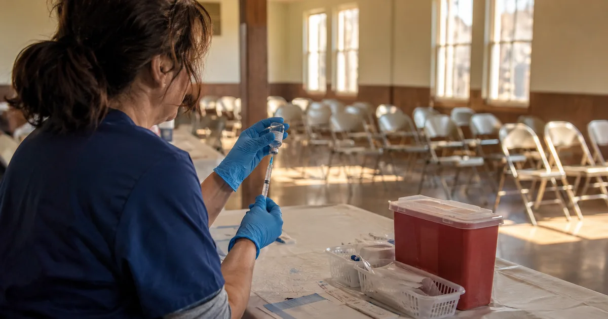 A public health nurse in navy scrubs and blue gloves draws a vaccine dose from a glass vial at a paper-covered folding table, with rows of empty folding chairs behind her in a sunlit community hall