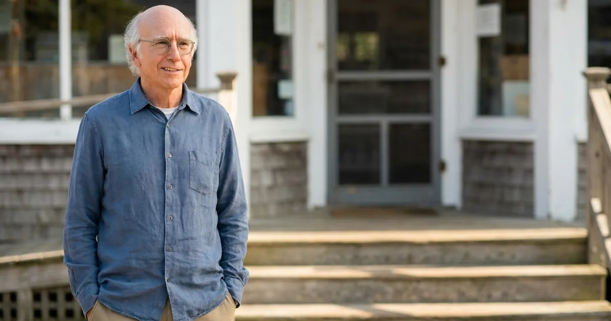 Larry David in a blue shirt standing with hands in pockets outside a weathered clapboard general store, smiling faintly and looking off to one side