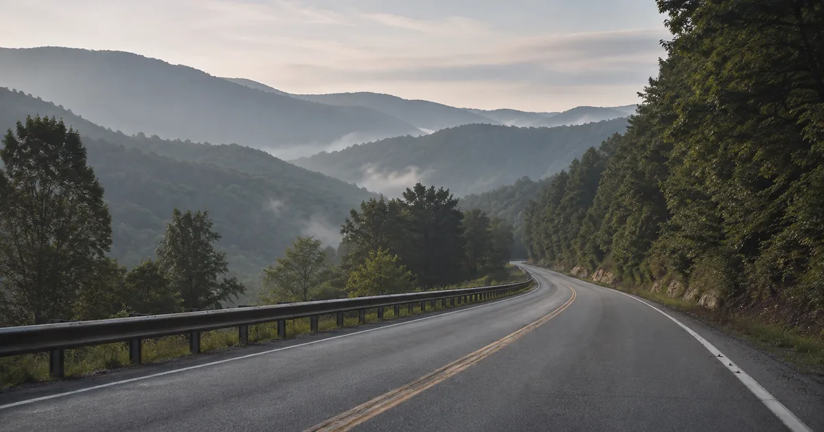 An empty two-lane rural highway curves through fog-filled Appalachian ridgelines at dawn, a metal guardrail running along the left shoulder and a narrow paved shoulder on the right