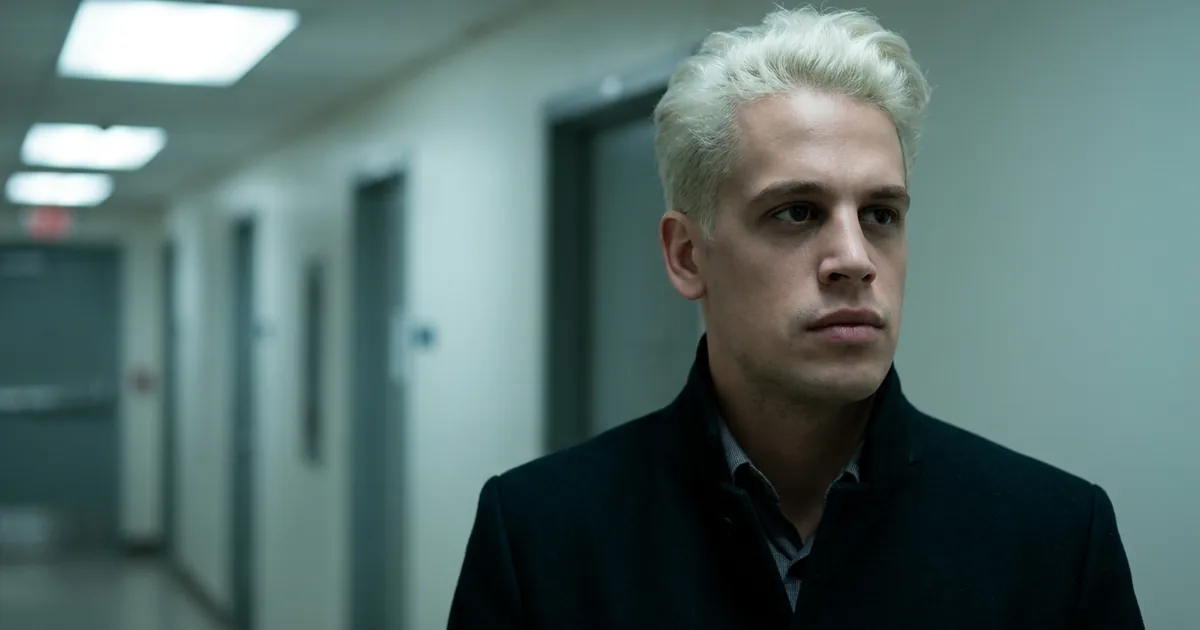 Milo Yiannopoulos, bleached-blond hair and a dark jacket, standing unsmiling in a pale fluorescent-lit government building corridor