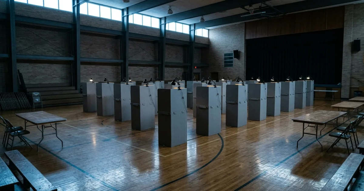 Rows of grey voting privacy booths with small lamps stand on a wooden gymnasium floor in cold morning light