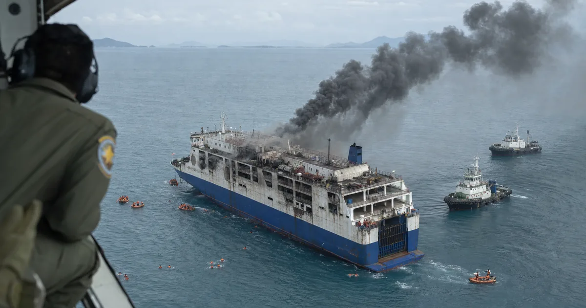 Aerial view from a rescue aircraft of a scorched blue and white roll-on roll-off ferry adrift at sea, black smoke pouring from its upper decks, with tugboats and small orange inflatable boats alongside and people in life jackets in the water
