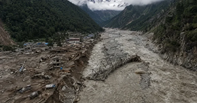 Aerial view of a Himalayan river valley after a flash flood, with a collapsed steel truss bridge lying in a churning grey torrent and a mountain village reduced to mud, splintered timber and buried vehicles