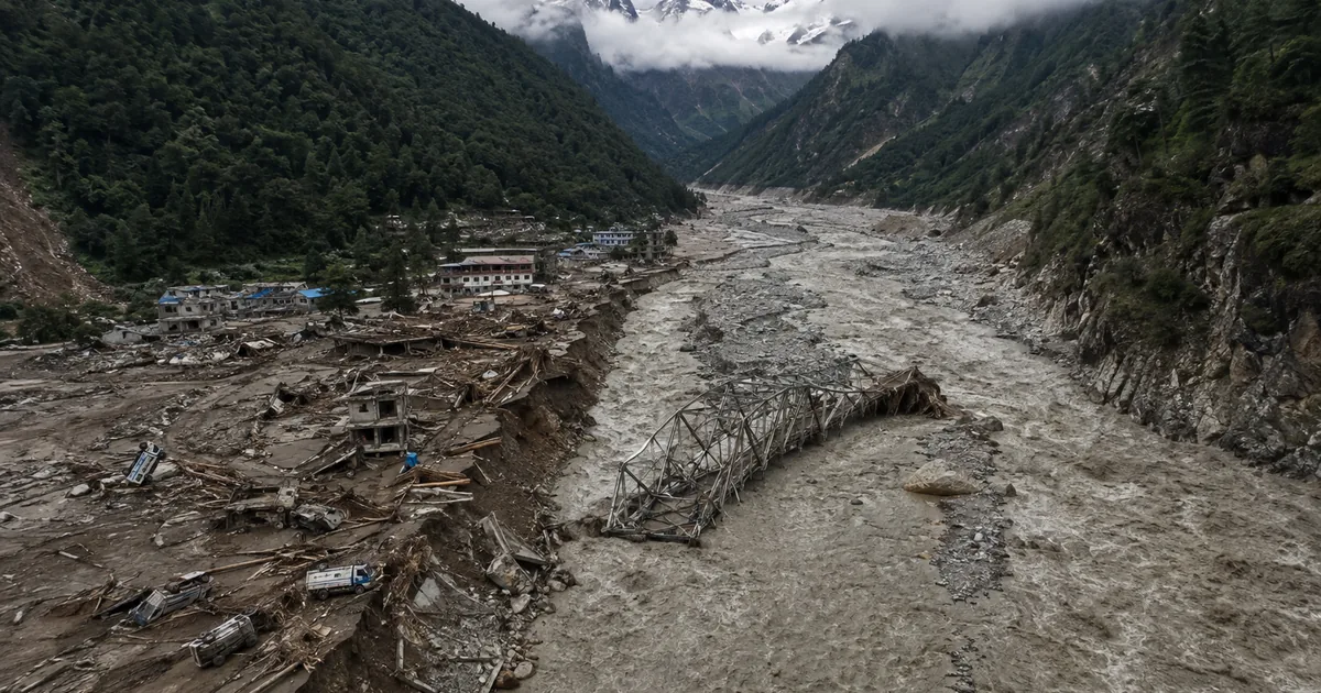 Aerial view of a Himalayan river valley after a flash flood, with a collapsed steel truss bridge lying in a churning grey torrent and a mountain village reduced to mud, splintered timber and buried vehicles