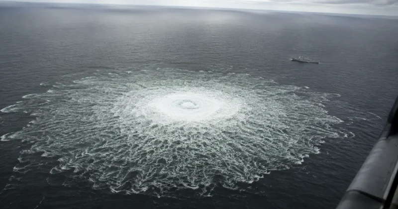 Aerial view of a wide circular patch of churning white water on the grey Baltic Sea where gas boils to the surface, with a naval patrol vessel standing off in the distance