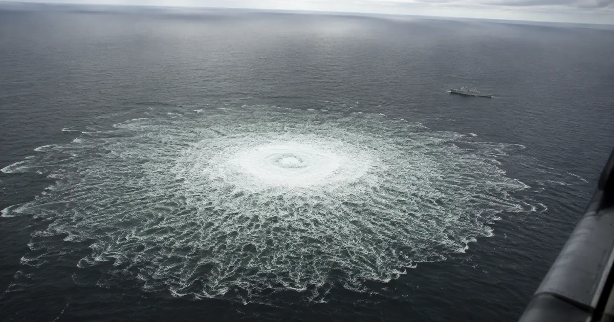Aerial view of a wide circular patch of churning white water on the grey Baltic Sea where gas boils to the surface, with a naval patrol vessel standing off in the distance