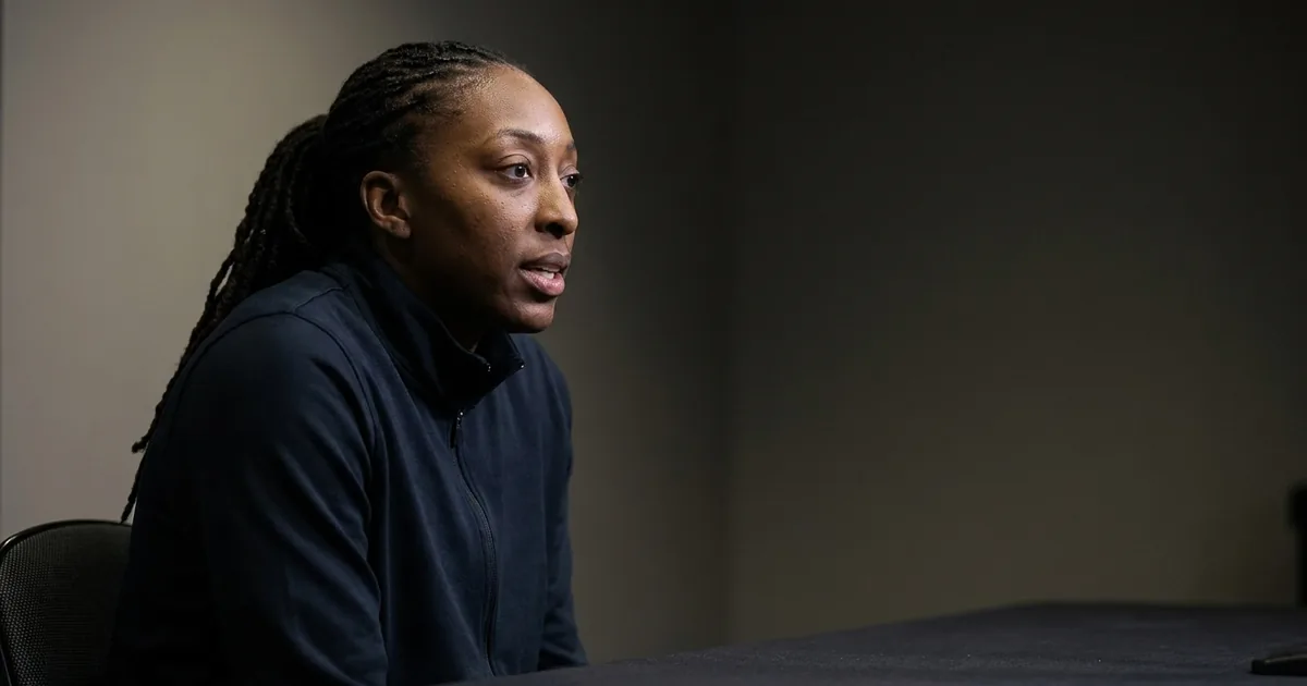 Nneka Ogwumike, in a plain dark warmup jacket, speaking mid-sentence at a press table against a dark backdrop