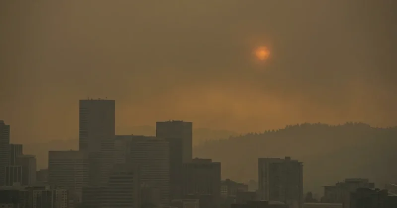 A Pacific Northwest city skyline reduced to grey silhouettes behind thick wildfire smoke with a dim orange sun disc burning through the haze