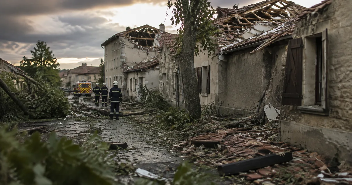 Firefighters on a debris-covered village street beside stone houses whose terracotta tile roofs have been torn away, under a heavy post-storm sky