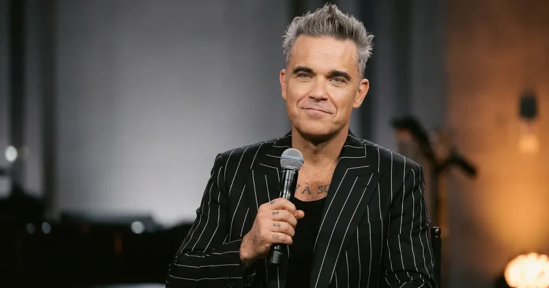 A greying pop singer in a dark pinstriped jacket holds a microphone mid-sentence with a wry half-smile