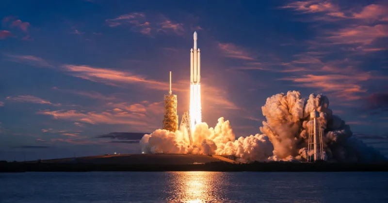 A three-core Falcon Heavy rocket climbing from a coastal launch pad at dawn, its exhaust plume lighting a huge steam cloud and reflecting off dark water