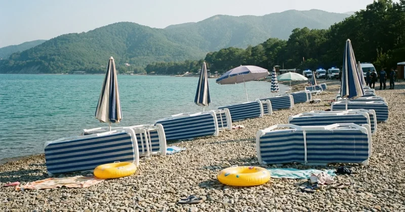 Abandoned striped sun loungers, tilted umbrellas and a child's inflatable ring left on an empty pebble beach beside turquoise water, with forested mountains behind and emergency vehicles with blue lights parked in the distance